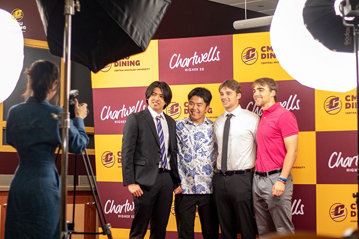 Students dressed in fancy clothes stand in front of a maroon and gold backdrop, posing for a professional group photo.