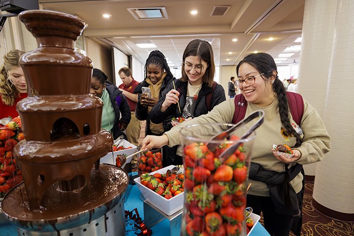 A small group of students smile while standing next to a table that has a chocolate fountain and strawberries on it.