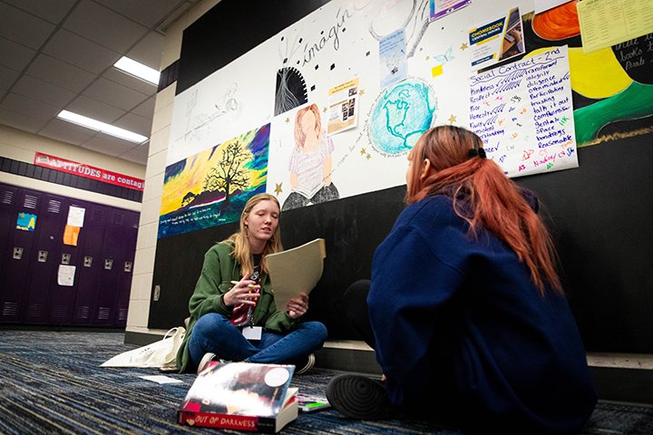In a carpeted hallway inside Mt. Pleasant High School, a CMU student and a high school student sit on the floor talking.