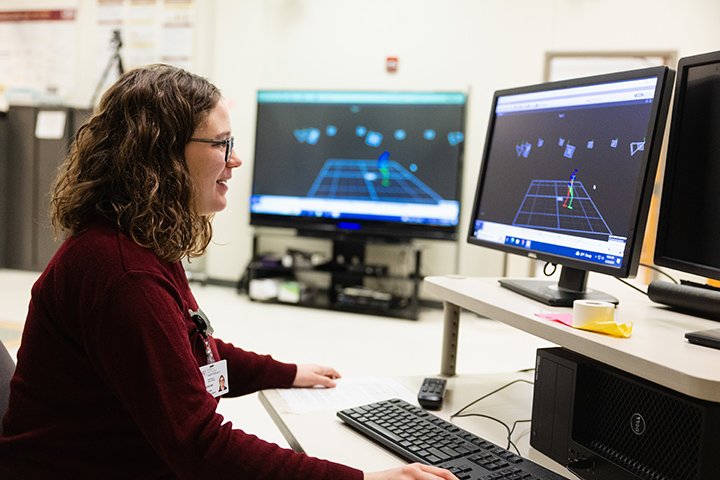 A female graduate student wearing glasses and a maroon long sleeve shirt sits at a computer with multiple screens.