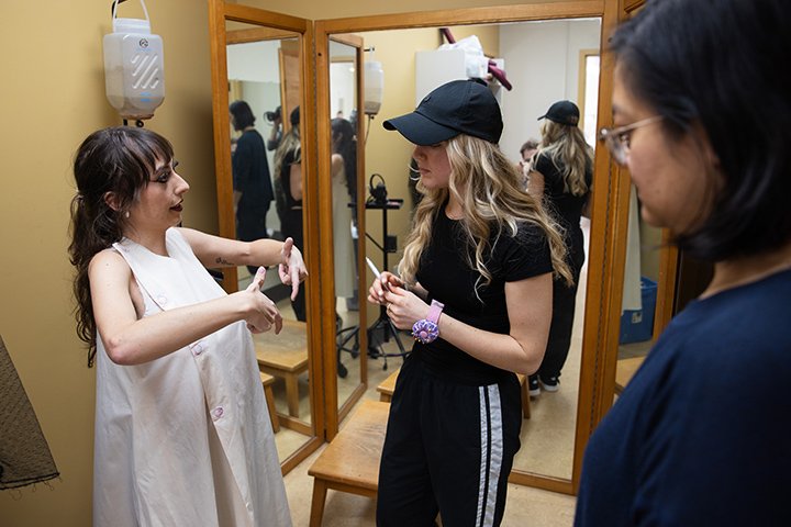 Two fashion design students, one wearing a white dress and the other wearing black pants, a black shirt and a black hat, stand in front of a mirror discussing clothing design while a third student in the foreground listens in.