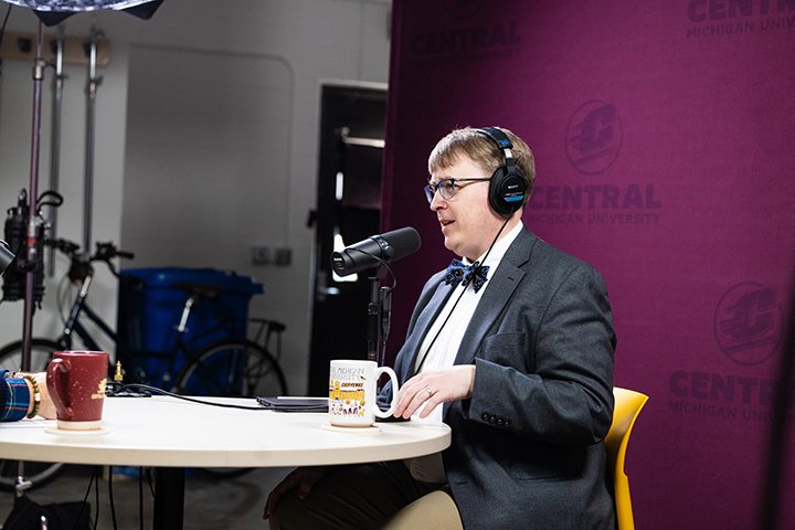 Wearing headphones, a suit coat, white shirt and signature bowtie, CMU physics professor Aaron LaCluyzé sits at a round table in front of a maroon background talking into a microphone during a podcast recording.