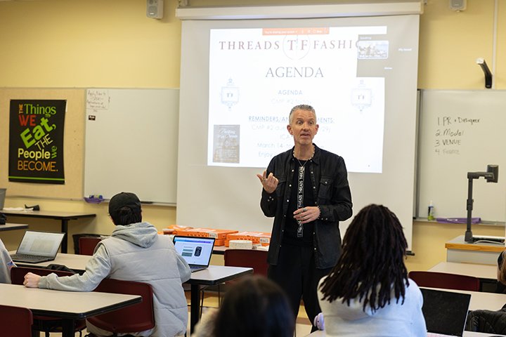 Wearing a black outfit, faculty member Ian Mull stands in front of a classroom of students as a projector behind him displays information about the Threads fashion show.