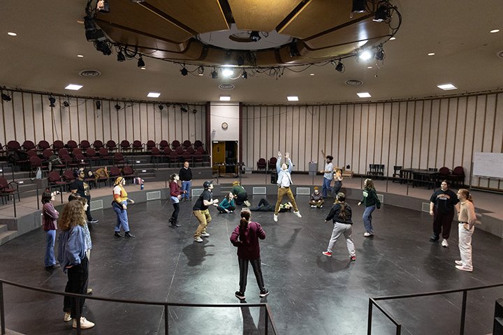 Approximately a dozen students stand in a large circle inside a large, round room taking part in an improv workshop with actor and CMU alum Larry Joe Campbell.