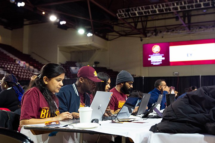 Three young people, one woman and two men, work on laptops on a table in a gymnasium.