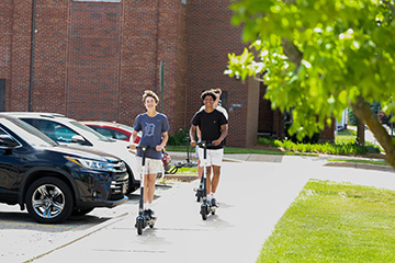 Three students wearing t-shirts and shorts ride electric scooters down the sidewalk next to a parking lot.