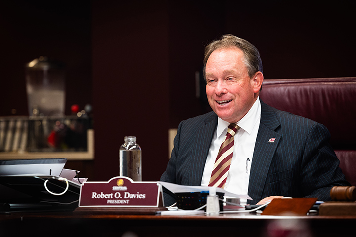 CMU President Bob Davies smiles white sitting at a large table during the Board of Trustees meeting.
