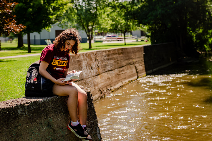 A female college student wearing a CMU shirt sits near a riverbank reading a book.