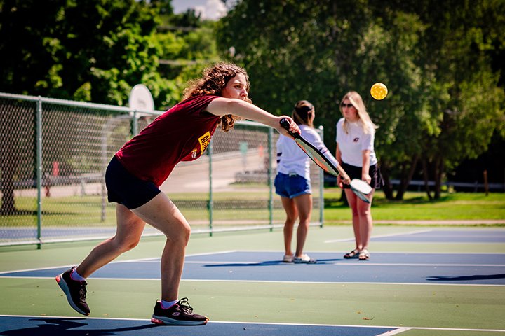 A CMU student in a maroon t-shirt plays on a pickle ball court as two people in white shirts stand on another court in the background.