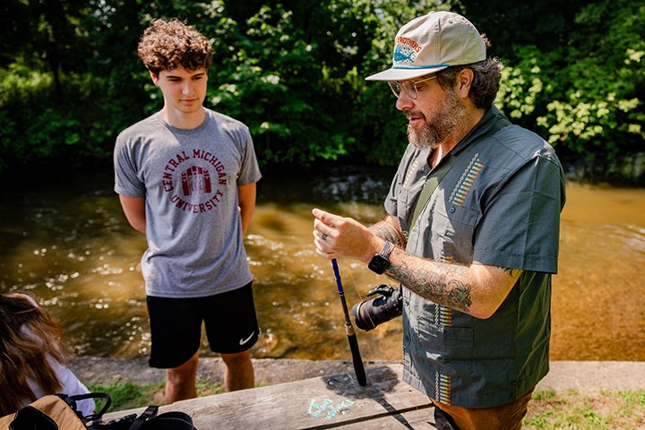 A bearded man in a hat and glasses prepares a fishing pole for a male CMU student.