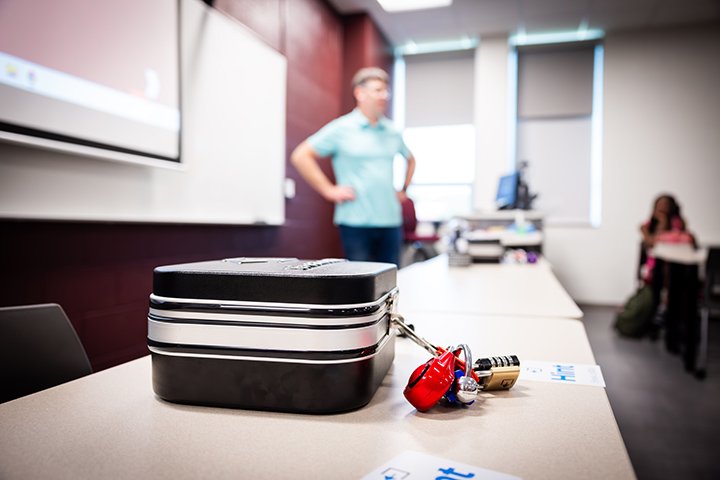 A locked suitcase sits on a table as a faculty member and students are blurred in the background.