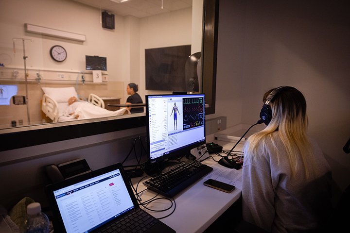 A person wearing a headset and looking at two monitors peers through a glass window into an exam room.