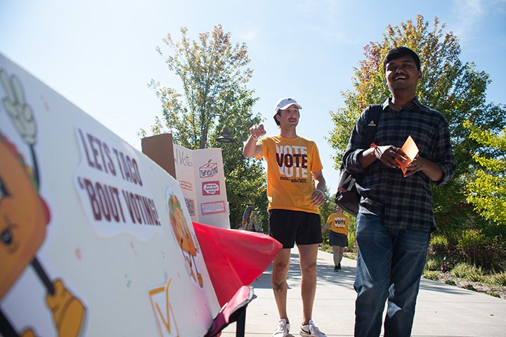 A CMU student in a shirt with the words Vote Vote Vote speaks to another student while standing next to a table.