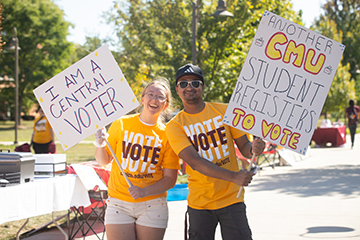 Two students wear "Vote Vote Vote" shirts while holding large, white signs encouraging people to register to vote.