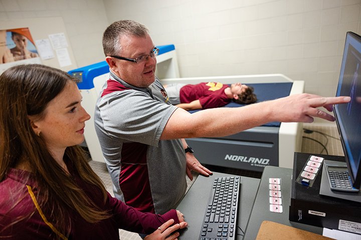 A male faculty member points at a computer monitor as a student looks on. In the background, another student lies on a body scanner.