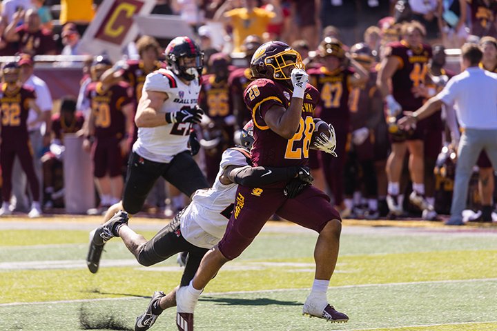 CMU football player B.J. Harris breaks a tackle while running down the field. In the background, players on CMU's sideline cheer.