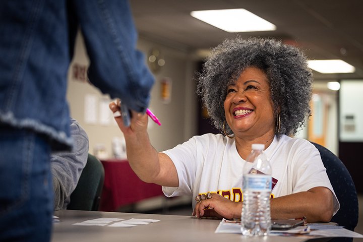 Trustee Regine Beauboeuf smiles while sitting at a table. She reaches out to hand a piece of paper to a student.