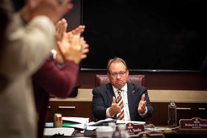 CMU President Bob Davies raises his hands in thanks towards a group of people standing and clapping for him.