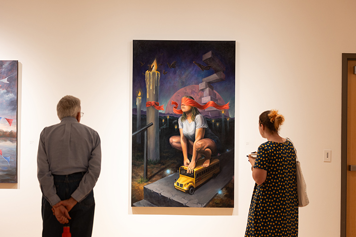 A view from behind a man and a woman who gaze at a painting of a blindfolded person sitting on a small school bus.