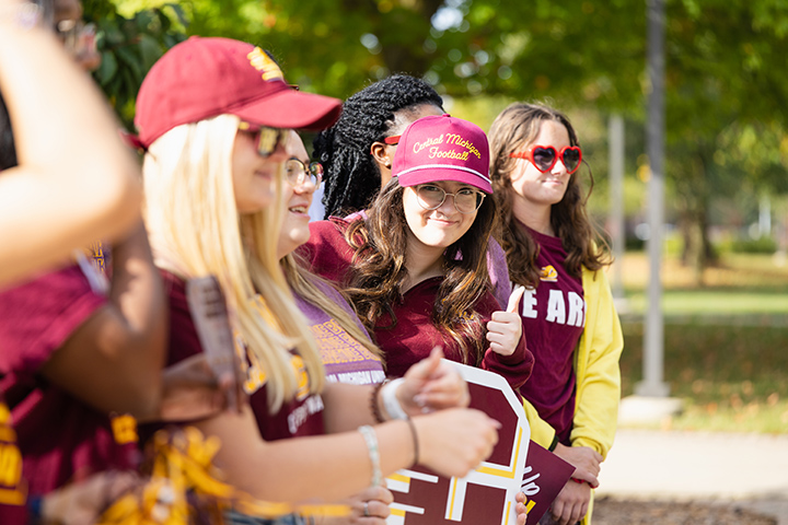 A student wearing a CMU football hat gives a smirk and a thumbs up while leaning out from a line of people.