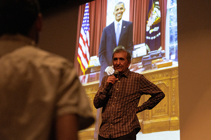 In a dark auditorium, Pete Souza stands on stage holding a microphone with an illuminated photo of President Obama on a large screen behind him.