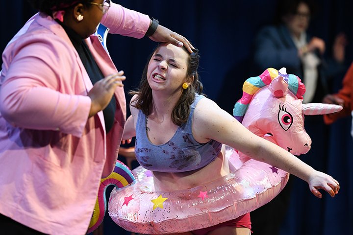 An angry looking young woman with a blow-up pink unicorn around her waist gets held at bay by her forehead by another young woman in a pink sweater.