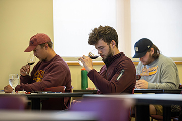 Three students sit at desks sniffing wine glasses filled with red wine.