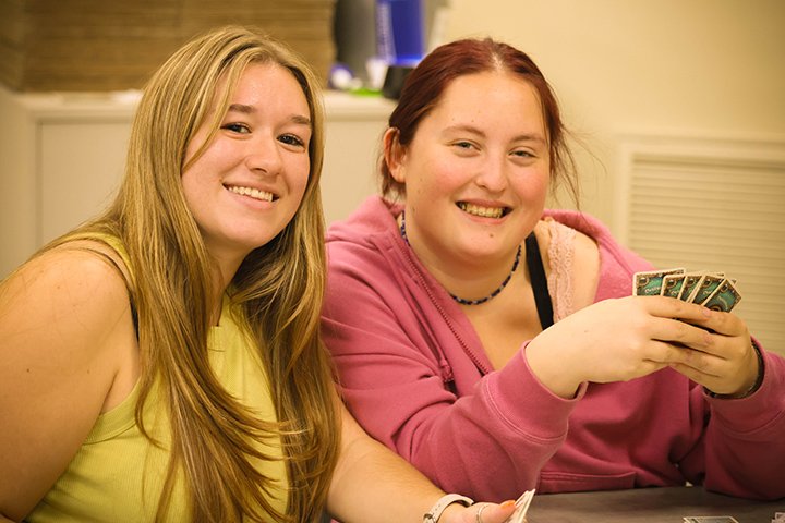 Two female college students sit at a table, holding playing cards and smiling at the camera.