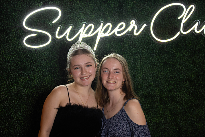 Two CMU students, one wearing a tiara, pose for a photo in front of a photo background that has the words Supper Club written in neon cursive letters.