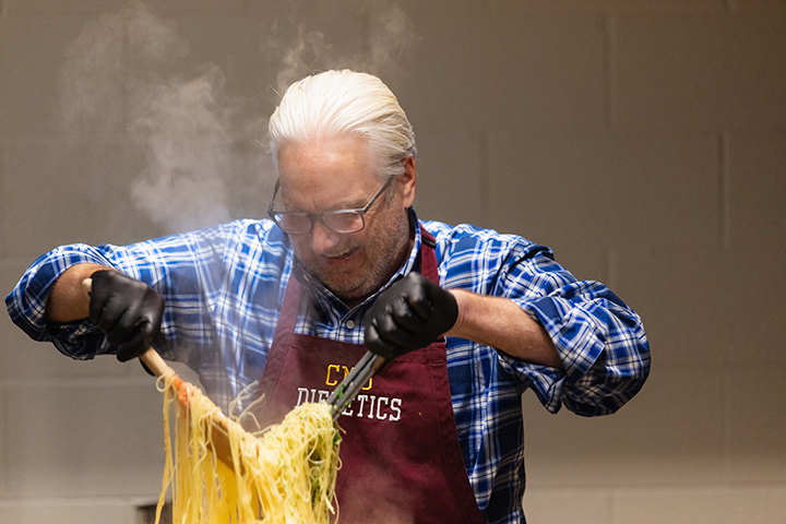 Jeff Fisher stands over a steaming pot of pasta, both hands holding large spoons with noodles hanging off of them.