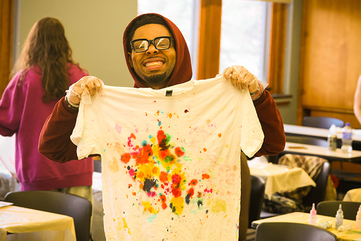 A student with glasses holds up a white tie dye t-shirt.