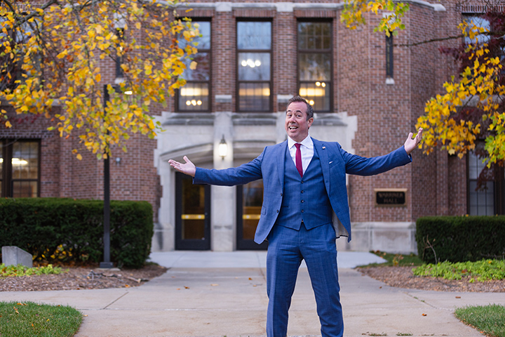 President Neil MacKinnon stands in front of Warriner Hall wearing a blue suit with his arms spread open.