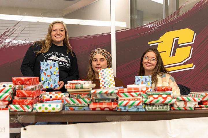 Three female college students sit at a table covered in books wrapped in wrapping paper.