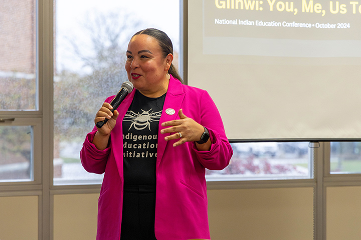 A woman in a pink sport coat stands in front of a room talking into a microphone.