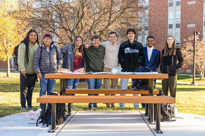 A group of eight students standing outdoors behind a picnic table.