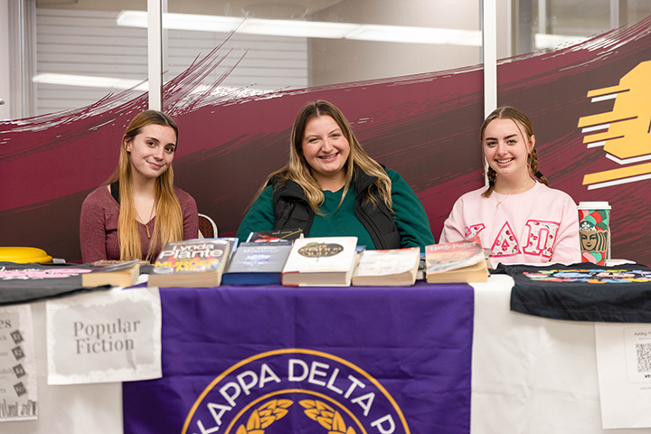 Three female students sit at a table covered in books and a sign that reads Popular Fiction.