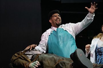 A scene from the musical Young Frankenstein depicts an actor dressed as Frankenstein's monster on a table and another actor yelling towards the sky.