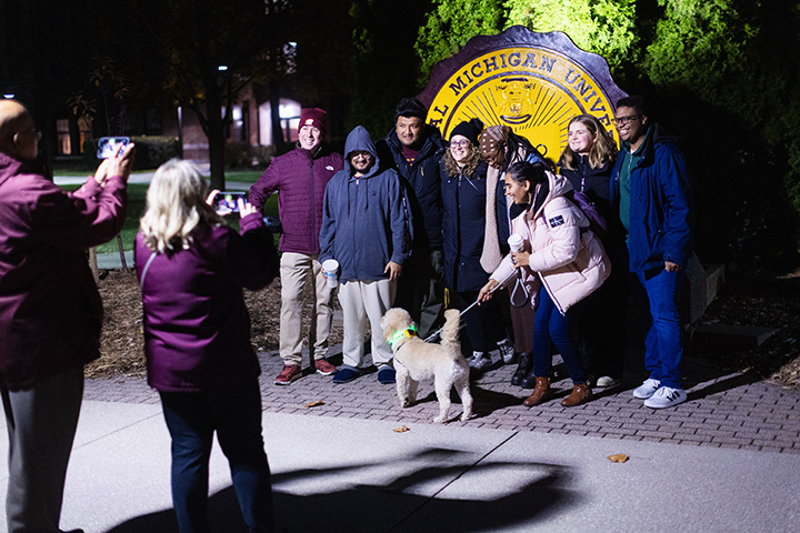 President MacKinnon, a dog and a group of students pose for a photo in front of the CMU seal at night.