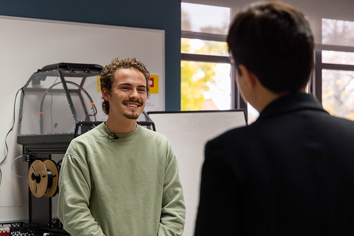 Two CMU students have a conversation while standing. One is wearing a light green sweatshirt. The other is wearing a black coat.