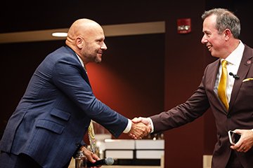 A man in a blue suit shakes hands across a table with a man in brown suit.