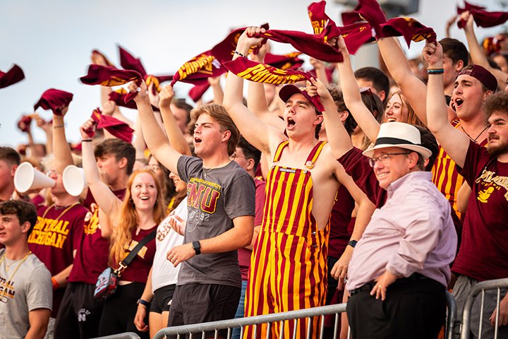 A crowd of CMU students cheer from the stands during a football game as President Davies stands nearby, gazing at the field.