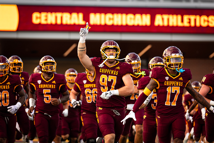 The CMU football team runs onto the field in a large group with a sign reading Central Michigan Athletics in the background.