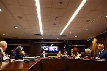 Eight people sit around a large wooden desk in a conference room.