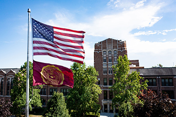 An American flag and a Central Michigan flag on a flagpole with trees and buildings in the background