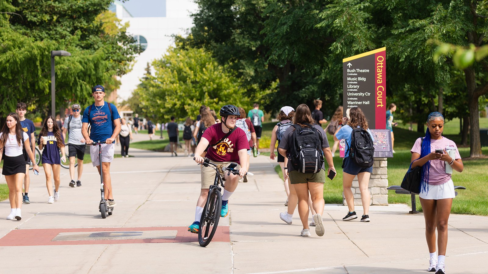 A busy group of students walk and ride bikes and scooters along a sidewalk. Large green trees flank the sidewalk.