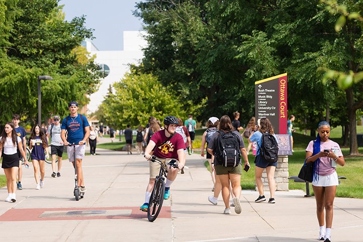 A busy group of students walk and ride bikes and scooters along a sidewalk. Large green trees flank the sidewalk.