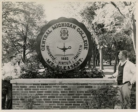 An undated black and white photo shows three CMU students standing next to the Central Michigan College seal.