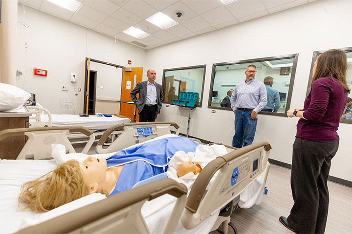 Three people look at a healthcare manikin lying on a hospital bed in a healthcare simulation room