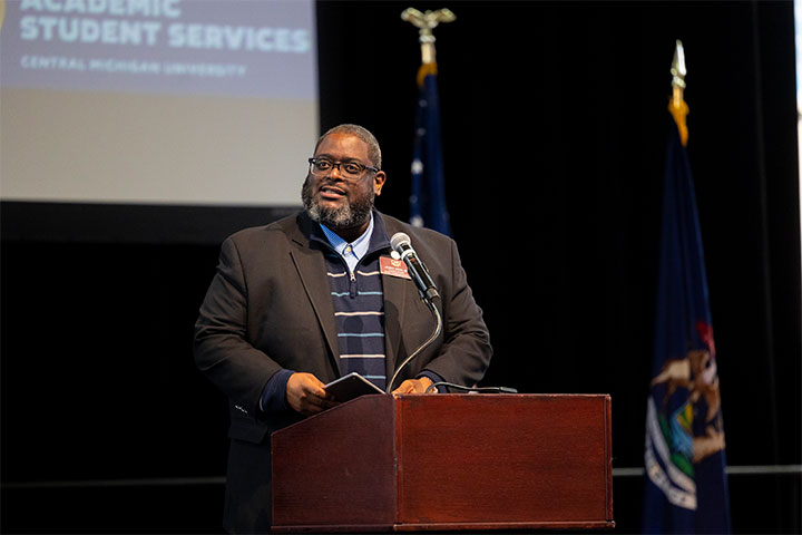 A Black man in a suit stands at a podium and reads from a book