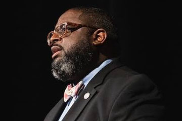 A close-up photo of a man wearing a dark suit and glasses.